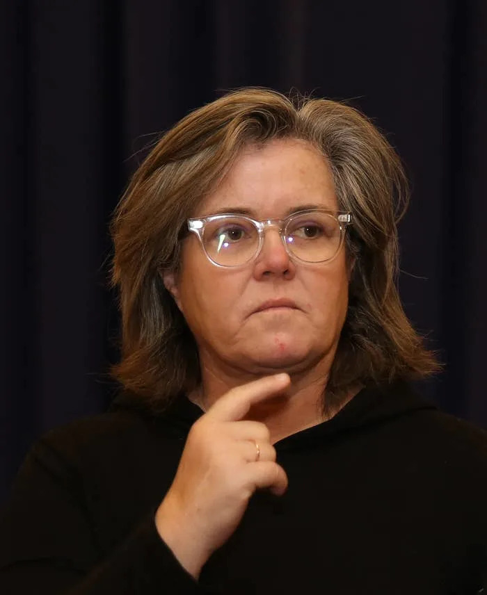 Rosie O'Donnell with medium-length hair and glasses looks thoughtful while touching her chin