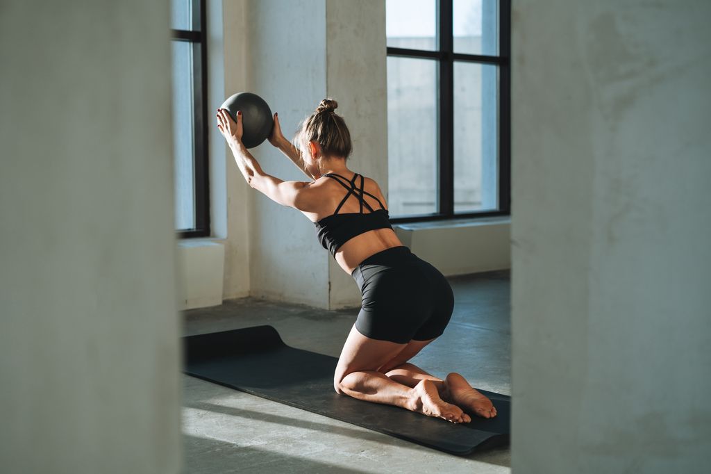 Young slim woman in sportswear doing Pilates stretching on mat with ball in modern studio