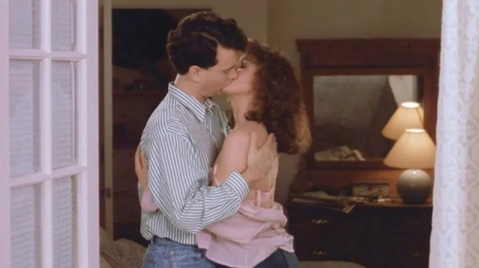 A couple embraces and kisses in a cozy room with a softly lit lamp and mirror in the background