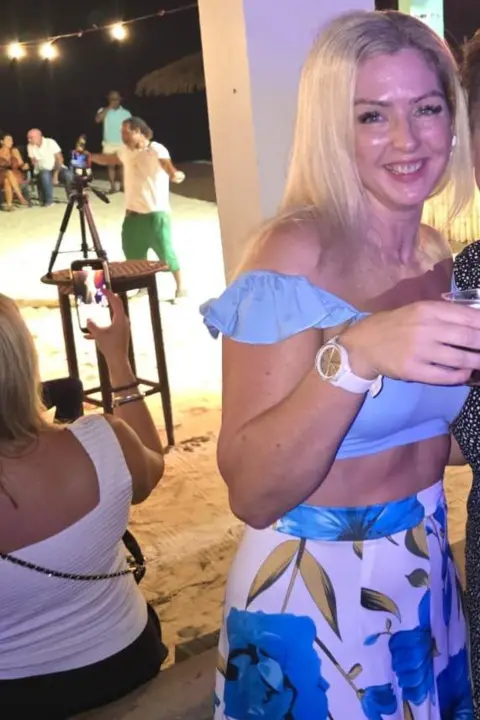 Cherie Wiles A women smiles at the camera and clinks aplastic cup of drink in a beach-side bar. She has long blonde hair and is wearing a sky blue off-shoulder top. Several men and women can be seen in the background. 