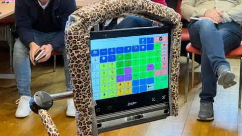 Close-up of an assistive communication device with a leopard-print frame displaying a colourful grid of symbols and letters. In the background, several people are seated on orange chairs in a room with wooden flooring.