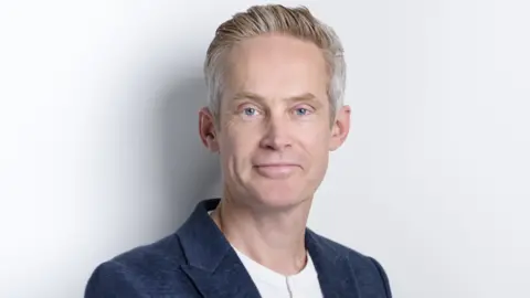 Guy Collis looking directly at the camera. He is stood against a white wall or background. He is wearing a blue suit jacket over a white t-shirt. 