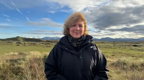 Portrait of Mannon Lewis, framed in the centre of the image. She has shoulder length wavey blonded or light brown hair and is smiling as she looks directly into the camera. She is wearing a black winter coat, with the white Natural Resources Wales logo in English and Welsh on the left side of the coast. She is wearing a scarf or neckwarmer, that is dark green and black. She is stood on pasture that is in fact sand dunes at Newborough Warren on Anglesey. Behind her can be seen the outline of mountains on the mainland, including the Carneddau and Eryri ranges, with some covered in snow. The sky is a mix of blue and some heavy dramatic clouds.