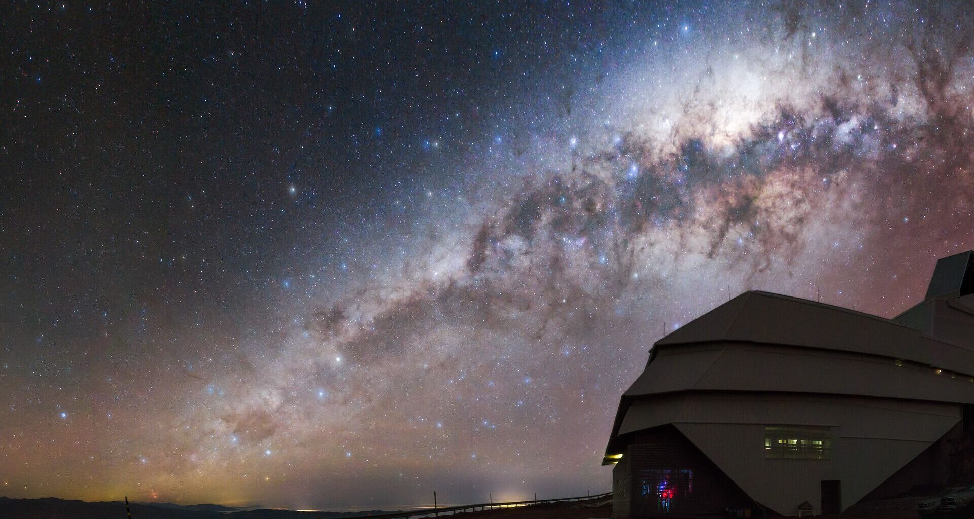 A glowing galaxy can be seen in the orange and blue night sky over the domed building of the Rubin Observatory