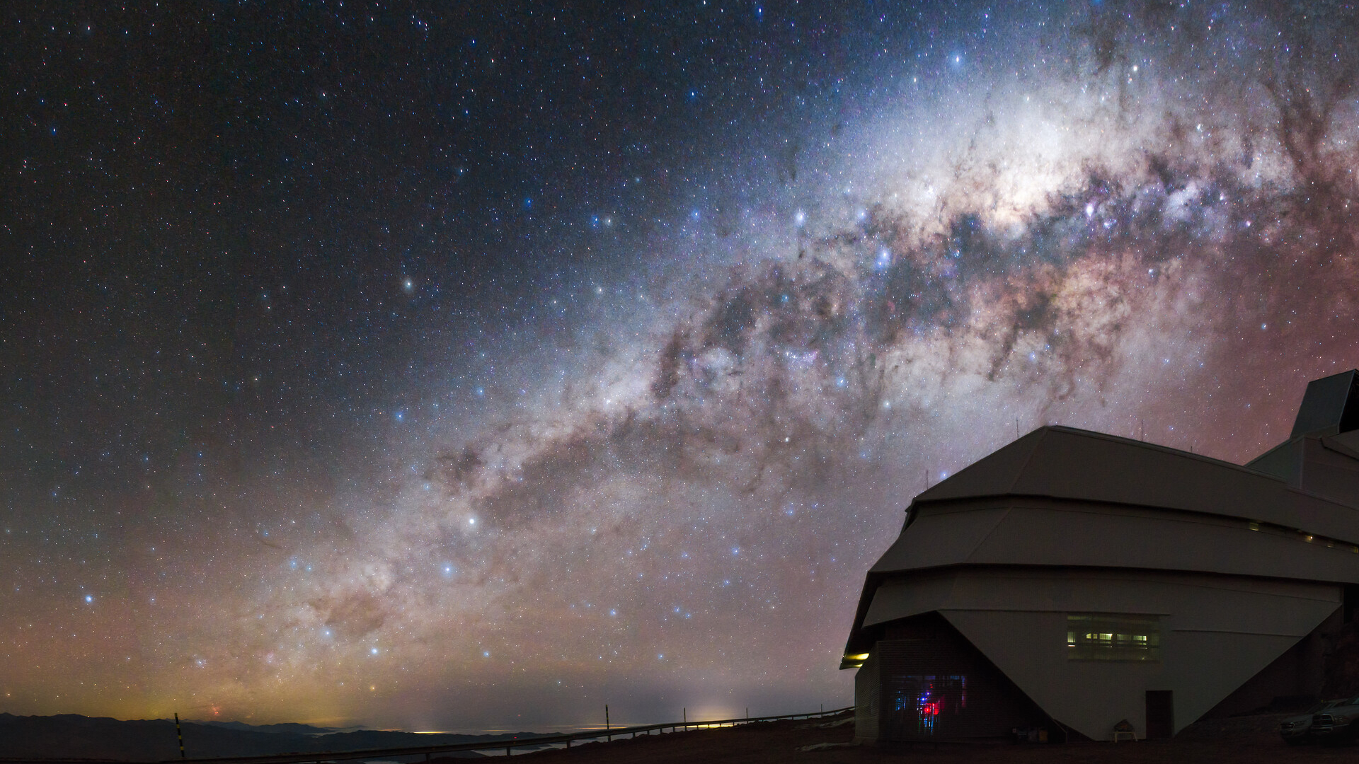 A glowing galaxy can be seen in the orange and blue night sky over the domed building of the Rubin Observatory