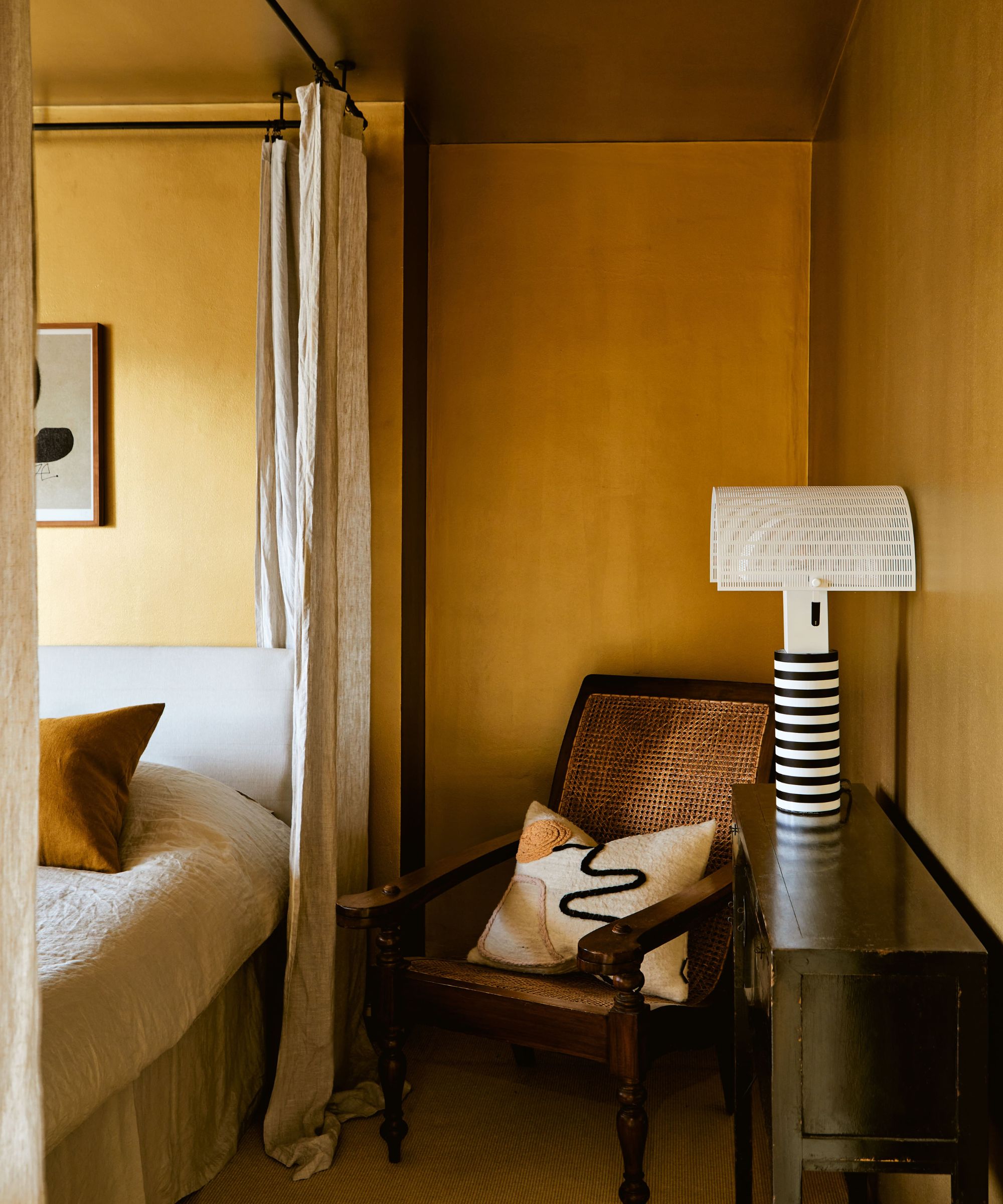 deep ochre yellow color drenched bedroom with a four poster metal bed, neutral bed linens and antique rattan and wooden chair