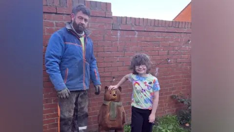 BBC A young girl with short wavy brown hair is wearing glasses and a multi-coloured T-shirt and has her hand on top of a small brown wooden bear with green scarf sculpture in a garden. A man stands on the other side of the bear. He has brown hair and a beard, and is wearing a blue waterproof jacket and black trousers with sawdust on them, as well as black gloves.
