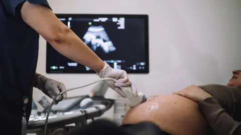 Getty Images Stock shot of a pregnant woman being given an ultrasound scan. You can see the lady's pregnant belly and a scanning tool being operated by a medical practitioner wearing gloves. Behind is an out-of-focus image of the baby appearing on screen.