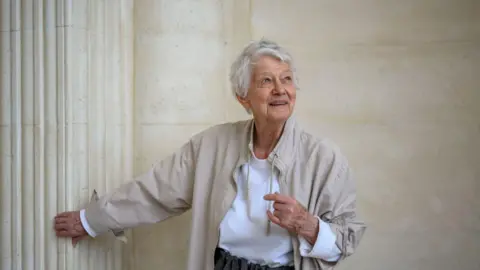 Getty Images Bridget Riley stood in a room with a light-coloured, rounded wall and a fluted pillar. She has an arm outstretched and is looking to the side of the frame.