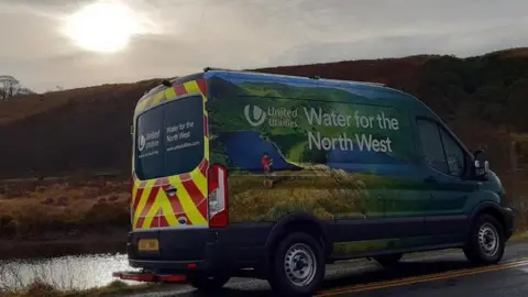 United Utilities A United Utilities van parked on a road in the countryside with a river the left and hills in the background. The sun is peering out through a cloud.