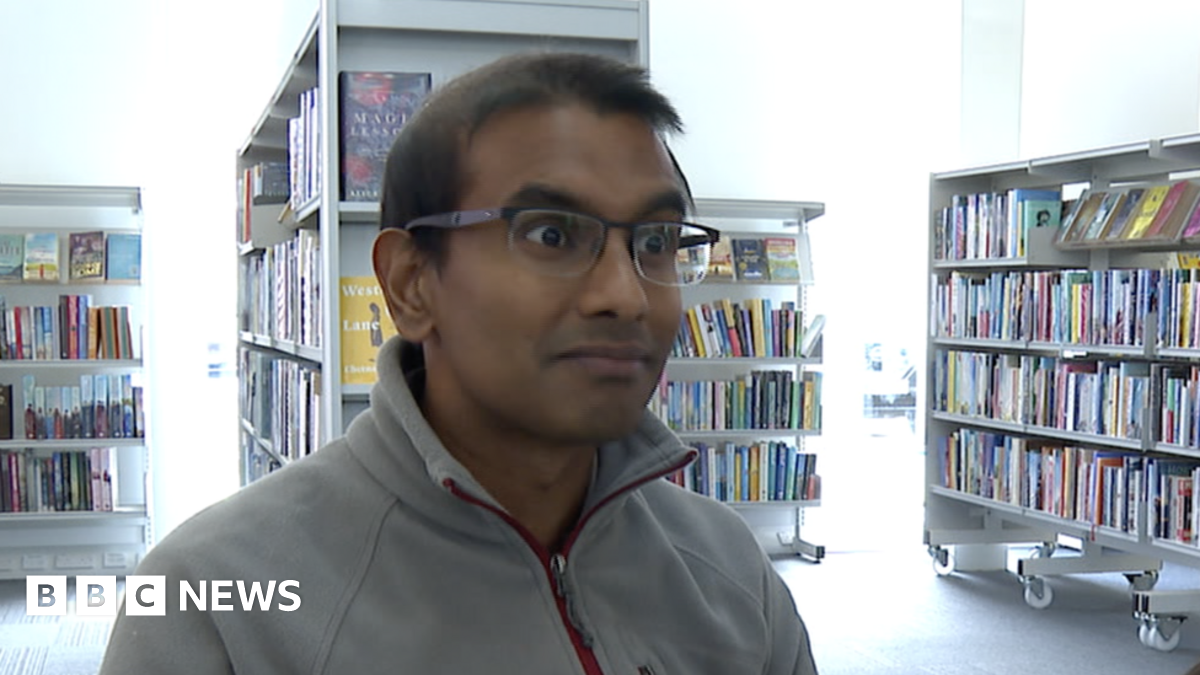 Doctor Kamlesh Sreekissoon is sat in a library with shelves filled with books behind him. He is wearing a fleece and glasses.