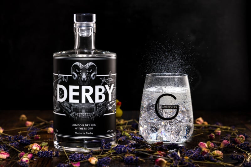 A bottle of Derby London Dry Gin stands next to a glass of gin and tonic with ice, surrounded by scattered dried flowers on a dark wooden surface with a black background.