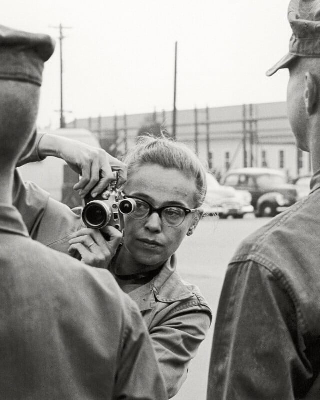 A woman with glasses and a camera takes a photo of two people in military uniforms outdoors, with cars and a building visible in the background.