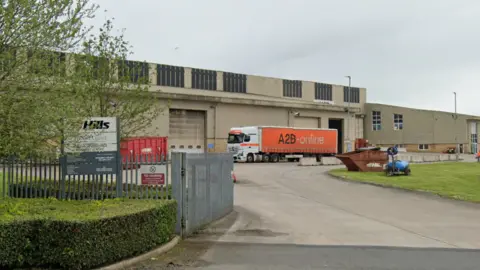 Google A view of the entrance into the waste recovery centre: Warehouse-type building with lorry outside. Large metal gates and fence. Tree and hedge outside. Sign of Hills waste at the front.
