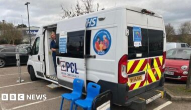 A woman in blue scrubs stands in a white medical vehicle with NHS logos and branding on it. The woman has blonde hair and smiles at the camera. The vehicle is parked in a car park with the door open and two blue plastic chairs in front of it.