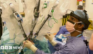 A surgeon in an operating theatre  wearing scrubs and surgical head wear. They are monitoring three robotic arms that are operating on a patients throat cancer.