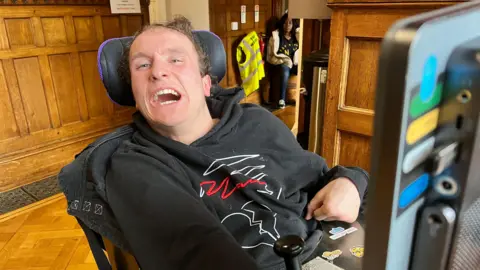 BBC A person seated in a powered wheelchair wearing a black hoodie with a white and red graphic design. A mounted communication device is visible in the foreground. The setting includes wooden panelled walls, a parquet floor, and a doorway in the background.
