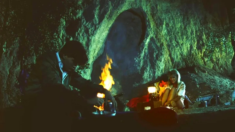 Charlie Sheen as Lance sitting with his head down in front of a campfire where Laura Dern as Tina and George Clooney as Ron are canoodling in Grizzly II: Revenge