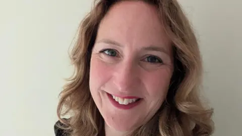 Claire Brown A woman with short curly hair and red lipstick is smiling. 