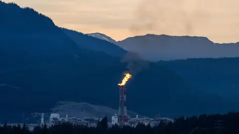 Bloomberg via Getty Images A white and red flare stack at sends fire up into the evening sky at the LNG Canada facility in Kitimat, British Columbia, Canada. In the background are mountains juxtaposed against a pale orange sky. 