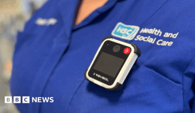 A group of five health care staff wearing uniforms stand in front of a hospital building. A man is at the centre of the image, the other four are women.