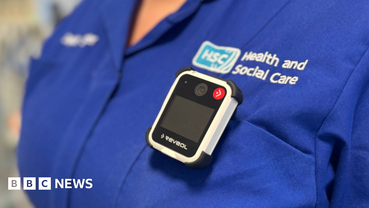 A group of five health care staff wearing uniforms stand in front of a hospital building. A man is at the centre of the image, the other four are women.