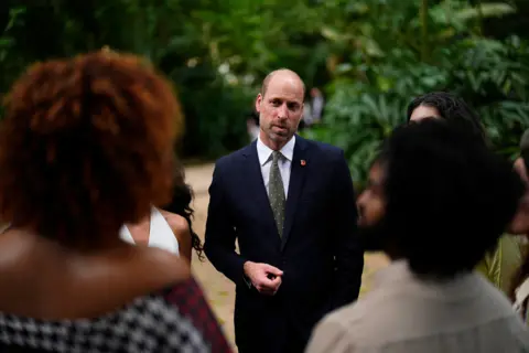 PA Media William, dressed in a dark suit, white shirt and tie, is facing camera as he stands on a path in the jungle looking quizzically at two people on either side of him with their back to the camera. 