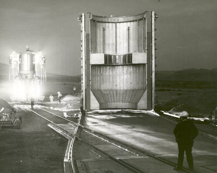 A black and white photo of a large, empty cylindrical structure, with a rocket releasing light in the background.