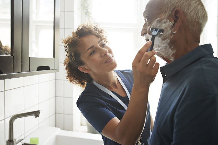 A nurse helps an older man shave.