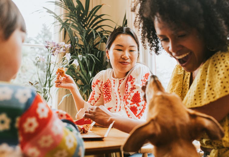 Three people sitting at a table eating and smiling at a dog asking for a bite