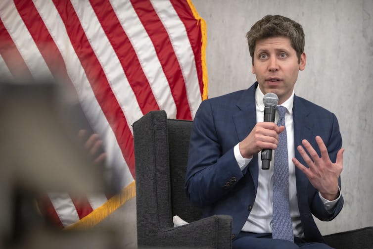 A middle-aged white man in a suit speaks into a microphone to an audience