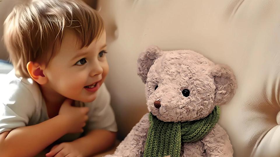 Marketing photo of a child grinning, looking at a teddy bear on a counch.