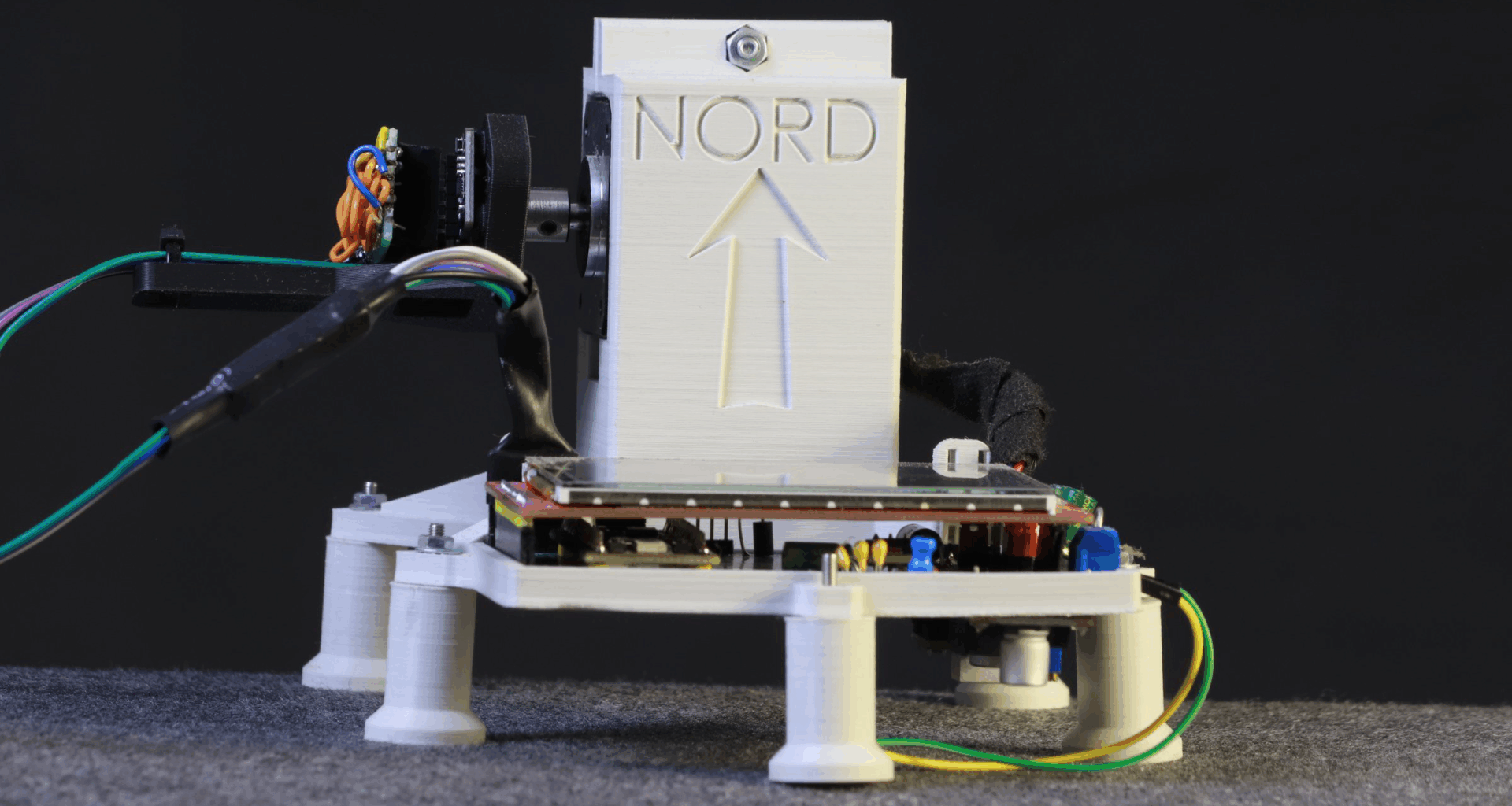 A 3D-printed assembly standing on short legs is visible. A portion extends upward with the word "Nord" sunk into it. Cables extend from one side of the upright portion, and a side view of a circuit board is visible at the front of the assembly.