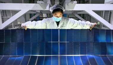 A worker in protective clothing and a mask inspects solar panel modules at a factory production line.