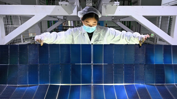 A worker in protective clothing and a mask inspects solar panel modules at a factory production line.