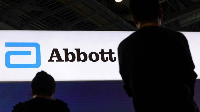 Two people stand in front of a large illuminated Abbott logo sign at a company booth.