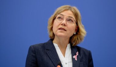 Henna Virkkunen speaks at a press conference, wearing a suit and a pink ribbon, in front of a blue background.