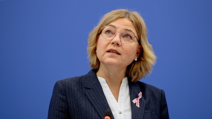 Henna Virkkunen speaks at a press conference, wearing a suit and a pink ribbon, in front of a blue background.