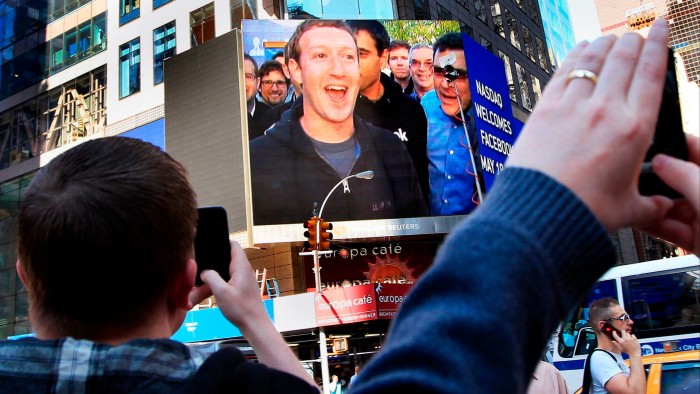 Mark Zuckerberg appears on a large screen in Times Square, smiling, as people take photos during Facebook’s IPO launch in 2012