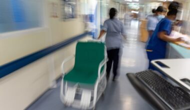 NHS staff in uniform work on a hospital ward; one staff member pushes an empty hospital chair down the corridor.