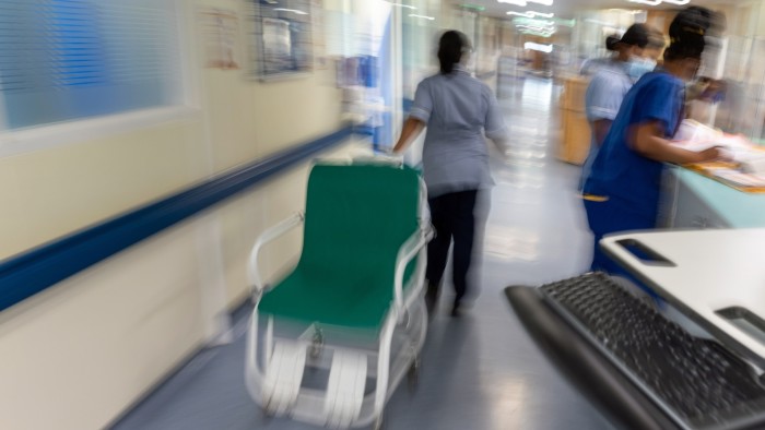 NHS staff in uniform work on a hospital ward; one staff member pushes an empty hospital chair down the corridor.