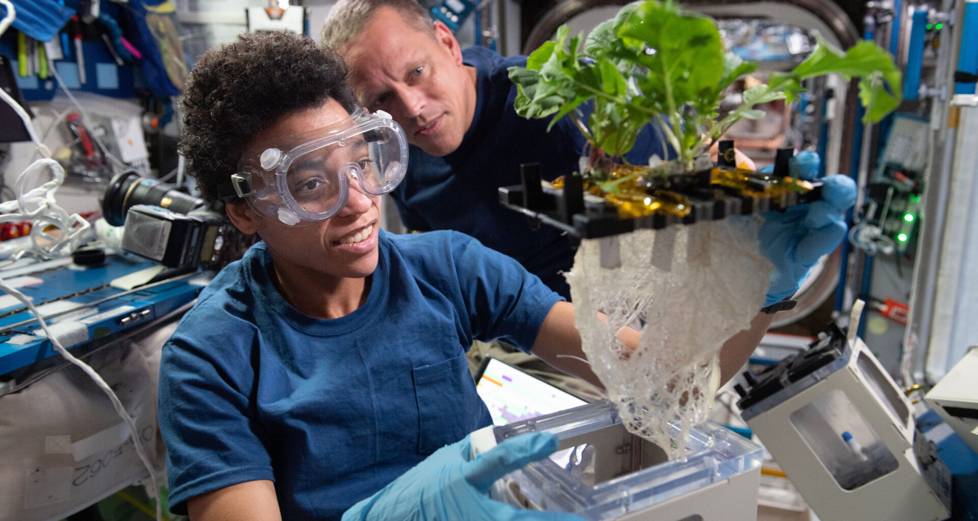 Jessica Watkins wears safety goggles and blue gloves while holding a plant growth unit with leafy greens and a large tangle of roots exposed. Bob Hines, in a black shirt, looks on from the background inside the space station laboratory.