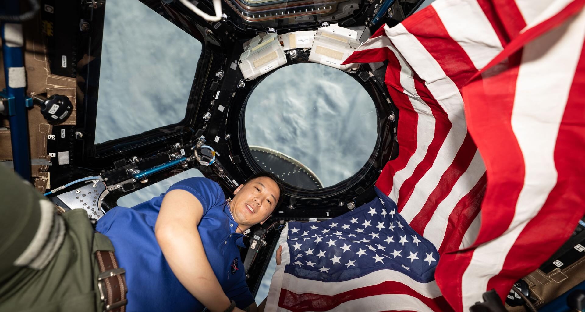 NASA astronaut and Expedition 73 Flight Engineer Jonnny Kim poses for a portrait with the American flag on the Fourth of July, or Independence Day, inside the International Space Station's "window to the world," the cupola. The orbital outpost was soaring 268 miles above a cloudy Atlantic Ocean at the time of this photograph.