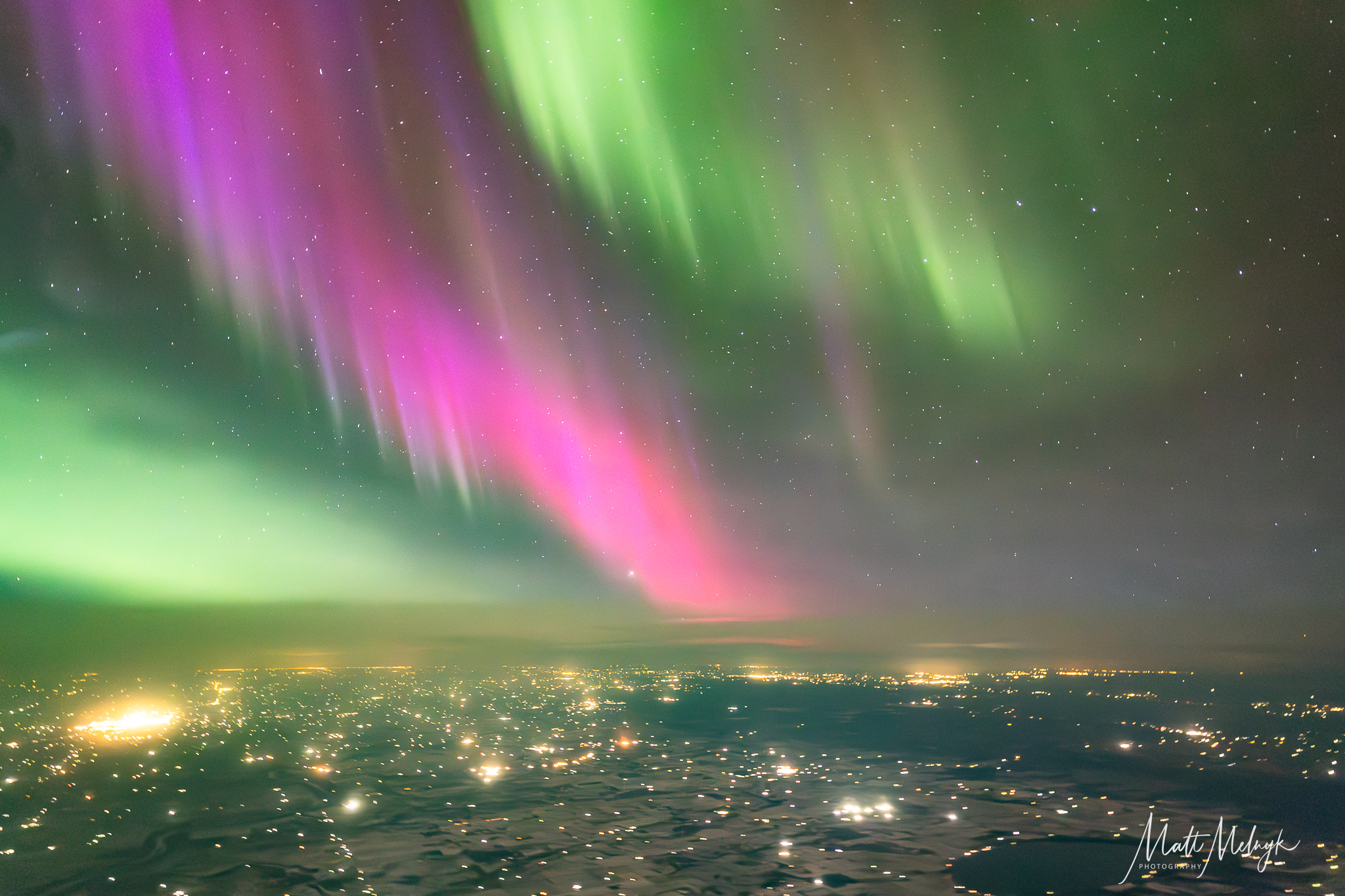 curtains of magenta and green light dance in the sky, yellow spots from city lights fill the scene below. 