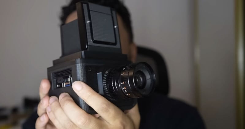 A person holds a large black camera with a prominent lens and a rectangular viewfinder, partially obscuring their face. The background is out of focus.
