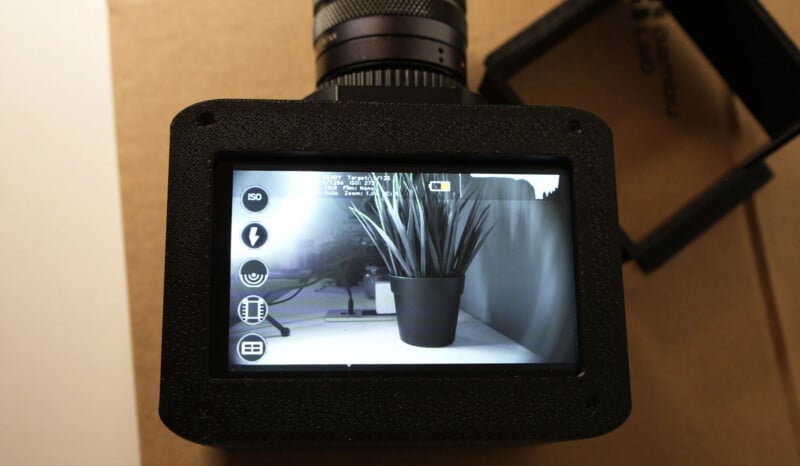 A close-up of a digital camera screen displaying a black potted plant on a table, with camera settings icons on the left side and technical details overlaid at the top of the display.