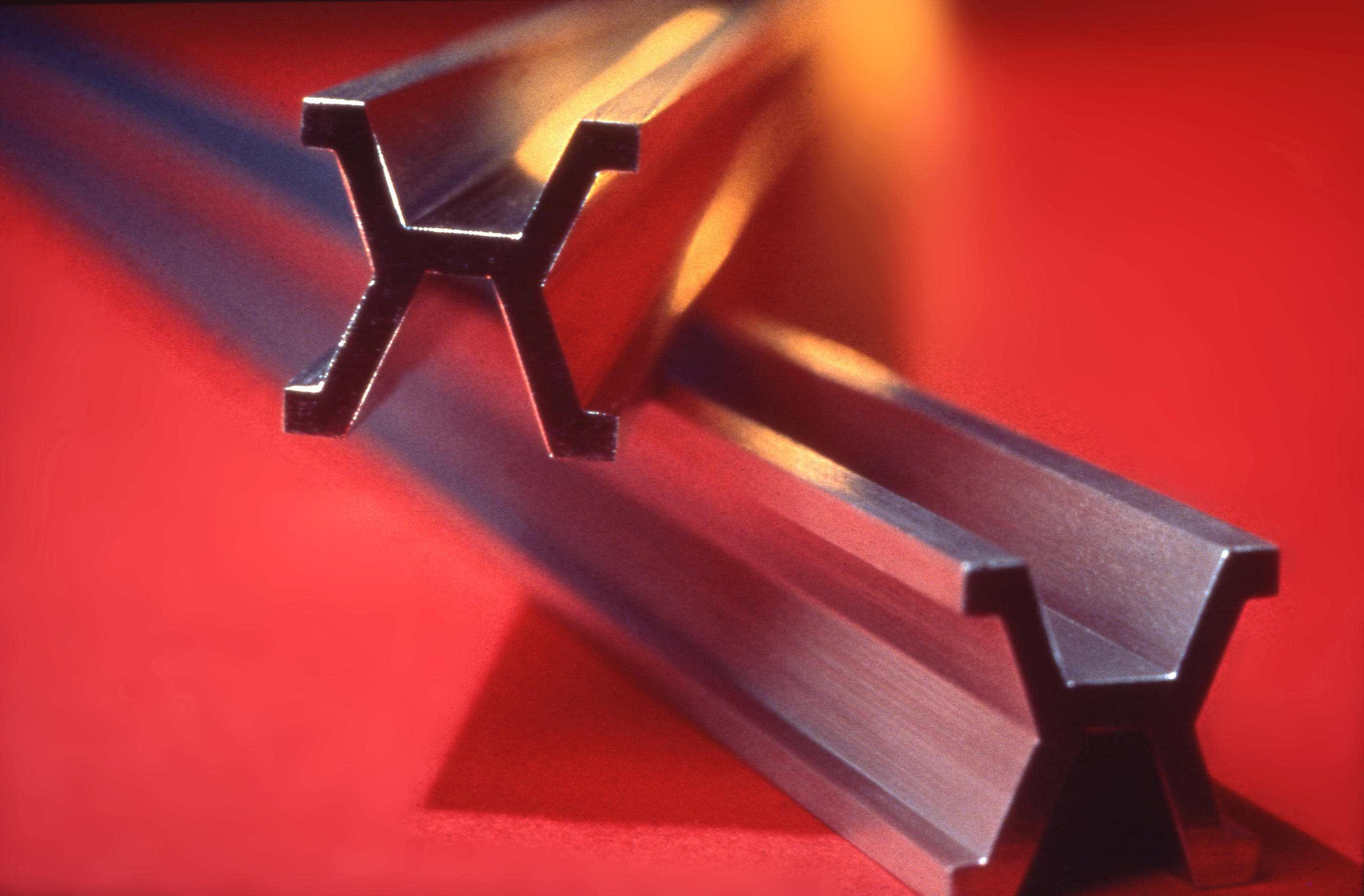 Close-up of two metal beams, each one meter long with an H-shaped cross-section, placed parallel on a red surface.