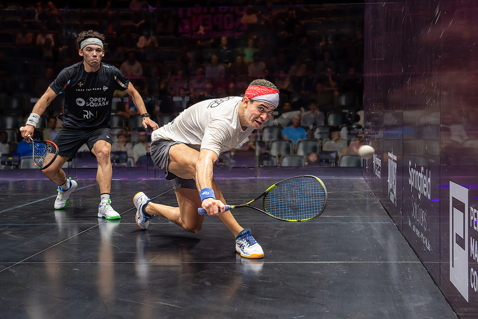 Two squash players compete intensely on a glass court, with one crouching low to hit the ball while the other readies for a return