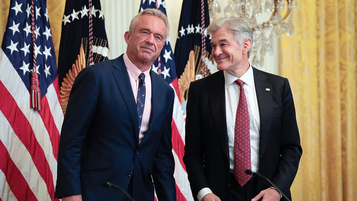 RFK Jr. and Dr. Mehmet Oz standing together in the East Room of the White House for event on health technology. Both seen smiling with American flags and other flags behind them.