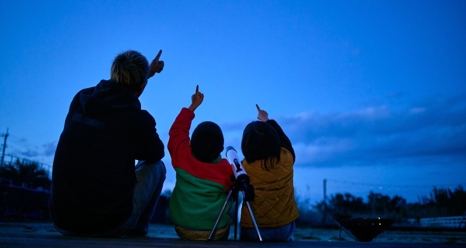 A man and a woman with a child between them all point at a night sky above them with a telescope on a tripod behind them